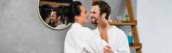 Femme gaie appliquant crème visage sur le nez du petit ami souriant en peignoir, bannière — Photo de stock