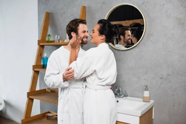 Mujer alegre aplicación de crema facial en la nariz de novio sonriente en albornoz - foto de stock
