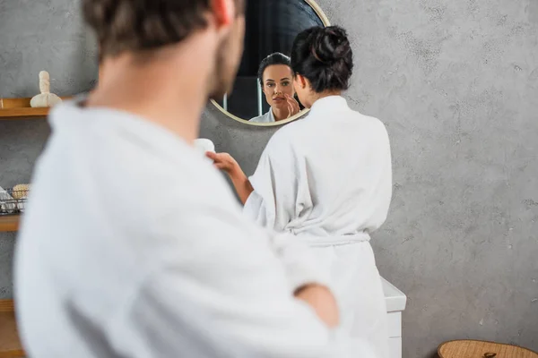 Frau trägt Gesichtscreme auf und blickt in Spiegel neben verschwommenem Freund im Vordergrund — Stockfoto