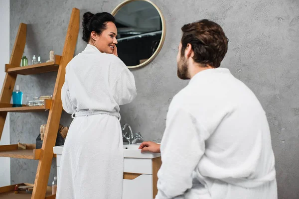 Mujer feliz en albornoz mirando novio en baño - foto de stock