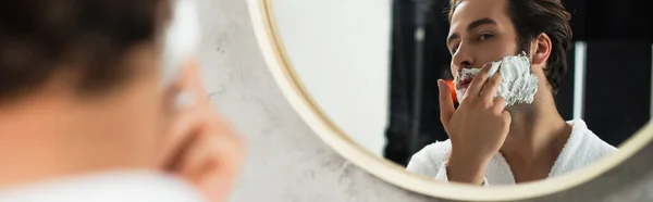 Человек наносит пену для бритья на лицо у зеркала, баннер — стоковое фото