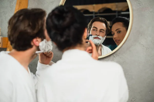 Spiegelung im Spiegel der Frau, die den Mann beim Rasieren im Badezimmer beobachtet — Stockfoto