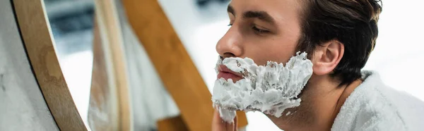Человек наносит крем для бритья на лицо у зеркала, баннер — стоковое фото