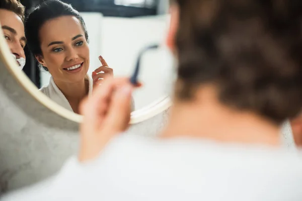 Spiegelung im Spiegel einer glücklichen Frau, die den Mann beim Rasieren im Badezimmer beobachtet — Stockfoto