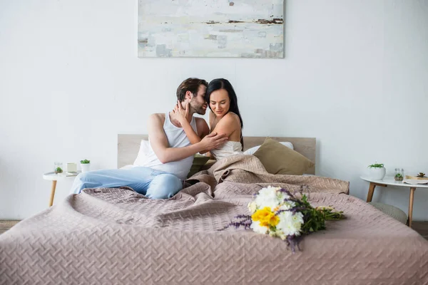 Buquê de flores na cama perto do casal satisfeito abraçando na cama — Fotografia de Stock