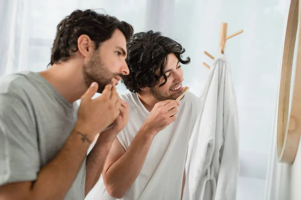 Blurred and tattooed gay man touching face while boyfriend brushing teeth and smiling in bathroom — Stock Photo