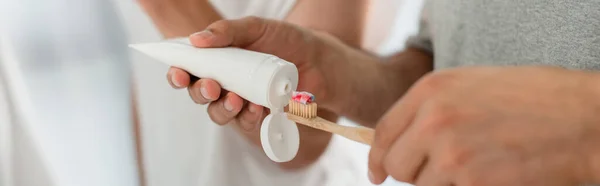 Vista recortada del hombre apretando la pasta de dientes en el cepillo de dientes cerca de novio en el baño, pancarta - foto de stock