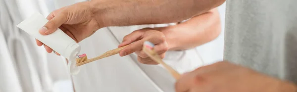 Vista parcial del hombre apretando la pasta de dientes en el cepillo de dientes cerca de novio en el baño, pancarta - foto de stock