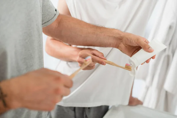 Teilbild eines Mannes, der im Badezimmer in der Nähe seines Freundes Zahnpasta auf die Zahnbürste drückt — Stockfoto