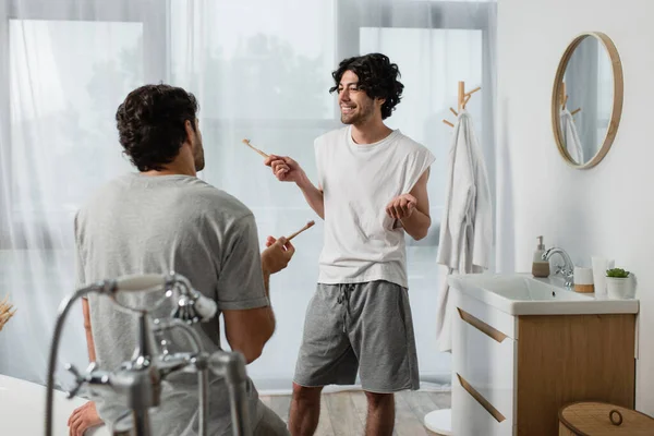 Barbudo gay pareja holding cepillos de dientes mientras hablando en cuarto de baño - foto de stock