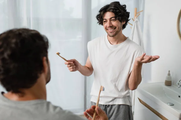 Barbudo gay hombre celebración cepillo de dientes mientras hablando con borrosa novio en cuarto de baño - foto de stock
