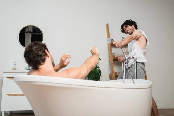 Glücklich Homosexuell Mann hält Duschkopf und zielt auf verschwommenen Freund in Badewanne — Stockfoto