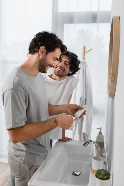 Feliz gay hombre mirando novio apretando dentífrico en cuarto de baño - foto de stock