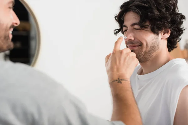 Uomo allegro e sfocato applicare la crema sul naso del fidanzato sorridente — Foto stock