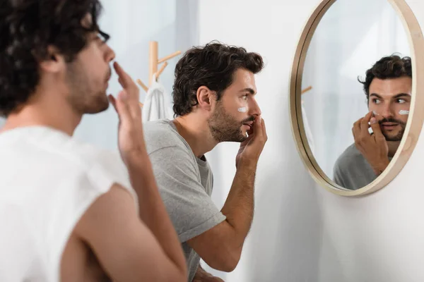 Homme appliquant de la crème sur le visage près de petit ami flou dans la salle de bain — Photo de stock