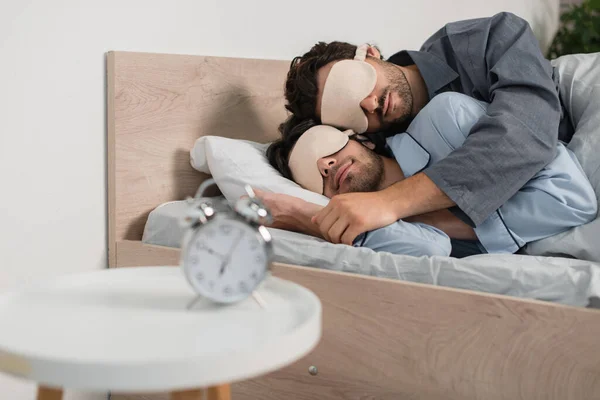 Gay couple dans les yeux masques câlins tandis que dormir dans lit près flou réveil sur chevet table — Photo de stock