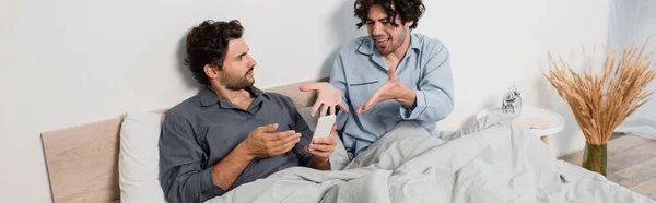 Unpleased man gesturing while looking at boyfriend chatting on smartphone, banner — стоковое фото