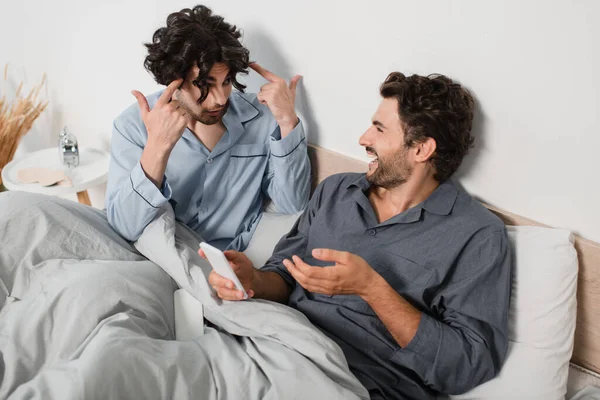 Displeased man quarrelling and gesturing while boyfriend holding smartphone and laughing in bedroom — Stock Photo