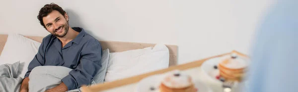 Verschwommener Mann hält Frühstückstablett mit Pfannkuchen neben glücklichem Freund im Bett, Banner — Stockfoto