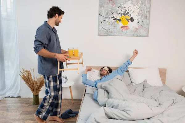 Man in eye mask stretching in bed near blurred boyfriend holding tray with orange juice — Stock Photo