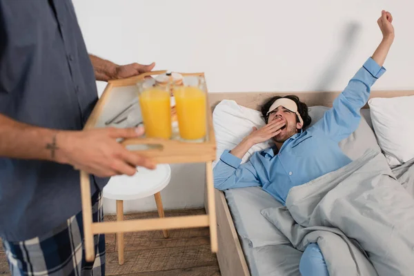 Homem em máscara ocular bocejando perto de namorado borrado segurando bandeja com suco de laranja — Fotografia de Stock
