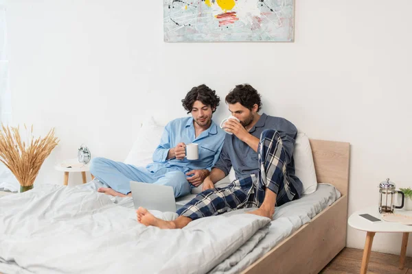 Гей пара пьет чай и смотрит кино на ноутбуке в постели — стоковое фото