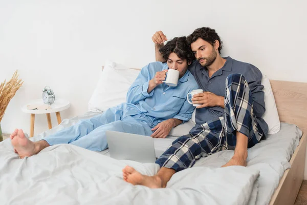 Gay pareja celebración tazas de té y viendo película en laptop en cama - foto de stock