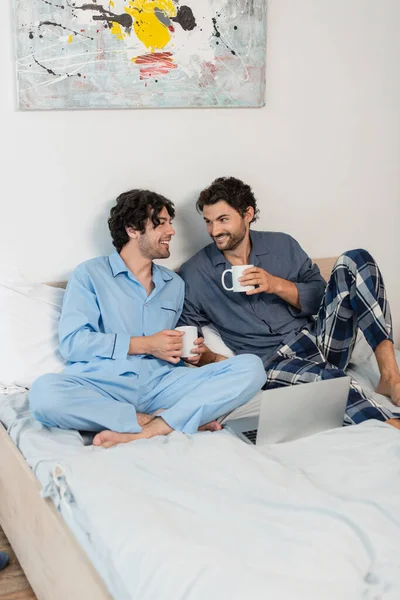 Felice coppia gay tenendo tazze di tè vicino laptop a letto — Foto stock