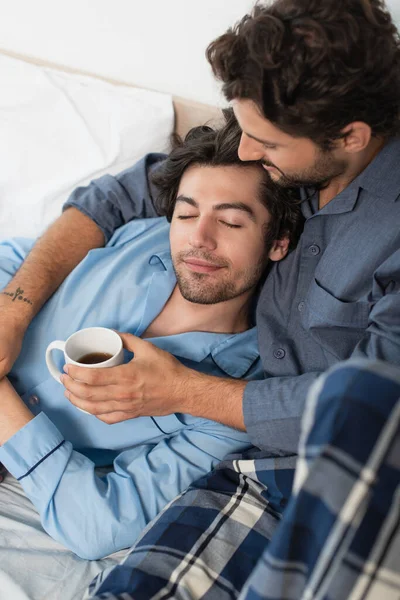 Tattooed gay man holding cup of tea and hugging boyfriend in bed — Stock Photo