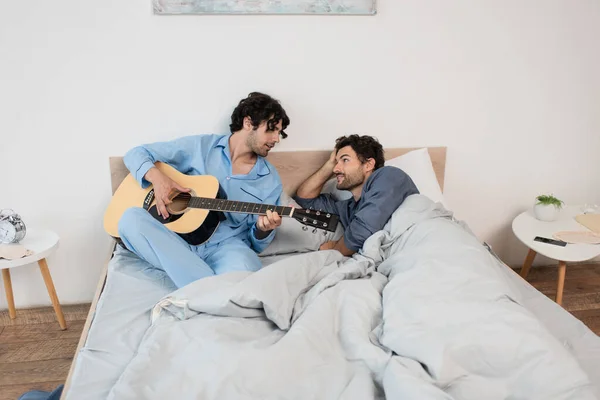 Gay uomo giocando acustica chitarra vicino fidanzato sdraiato su letto — Foto stock