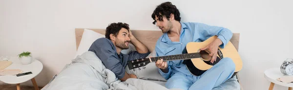 Gay uomo giocando acustica chitarra vicino fidanzato sdraiato su letto banner — Foto stock