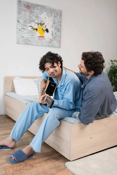Щасливий гей чоловік грає на акустичній гітарі біля хлопця на ліжку — стокове фото