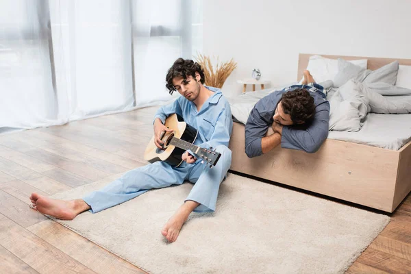 Gay hombre jugando acústica guitarra cerca novio en cama - foto de stock