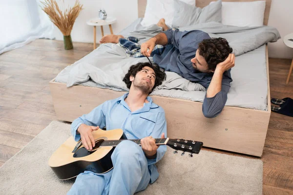 Gay uomo giocando acustica chitarra vicino premuroso fidanzato su letto — Foto stock