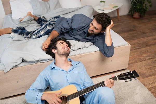 Улыбающийся гей играет на акустической гитаре рядом с бородатым парнем на кровати — стоковое фото