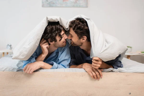 Vista lateral de feliz gay casal sob cobertor no quarto — Fotografia de Stock