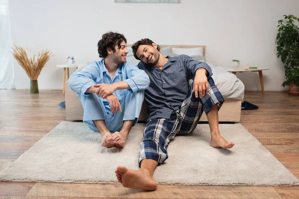 Alegre jovem gay casal sentado no tapete perto de cama — Fotografia de Stock