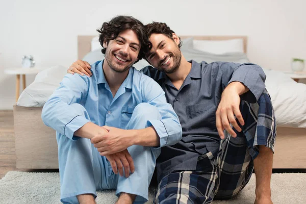 Alegre joven gay pareja sentado en alfombra cerca de cama - foto de stock