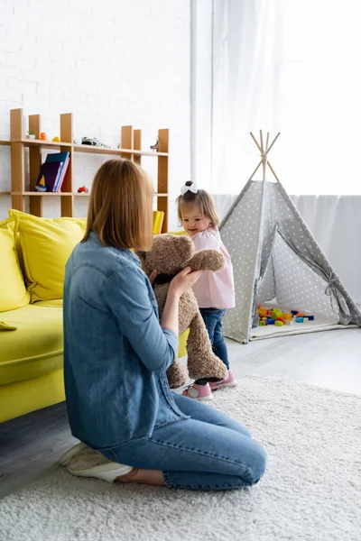 Maestra de jardín de infantes sosteniendo oso de peluche cerca de un niño con síndrome de Down en la sala de juegos - foto de stock