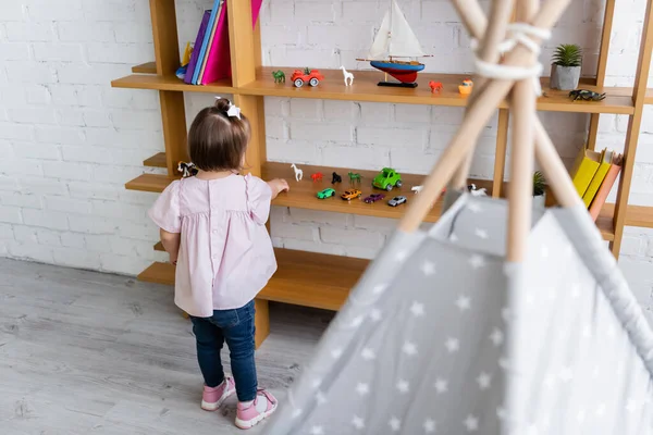 Vue arrière de la jeune fille avec le syndrome du duvet atteignant jouets sur étagère en bois — Photo de stock