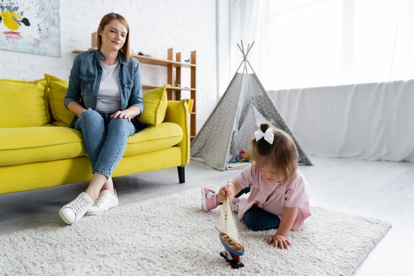 Tout-petit fille avec le syndrome du duvet jouer avec jouet bateau près flou maternelle enseignant assis sur canapé — Photo de stock