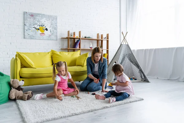 Kindergarten teacher sitting on carpet and playing with toddler girl with down syndrome near child in playroom — Stock Photo