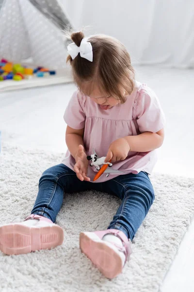 Niña con síndrome de Down sobresaliendo lengua y jugando con ovejas de juguete y avión en la alfombra - foto de stock