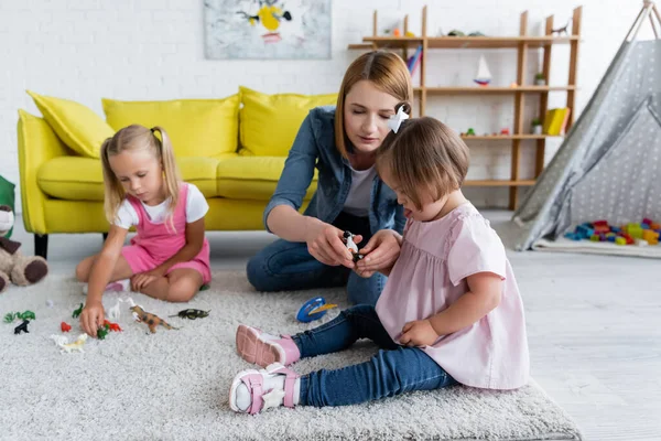 Maestra de jardín de infantes dando juguete a una niña con síndrome de Down cerca de un niño en edad preescolar borroso en la sala de juegos - foto de stock