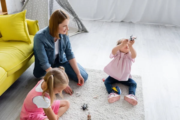 Vista ad alto angolo della maestra d'asilo guardando la bambina con la sindrome di Down che gioca con il giocattolo vicino al bambino in età prescolare offuscata nella stanza dei giochi — Foto stock