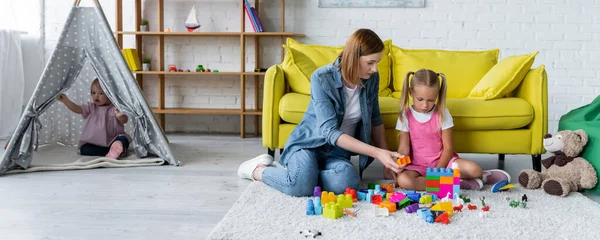 Kindergarten teacher playing building blocks with preschooler girl while kid with down syndrome sitting in tipi, banner — Stock Photo