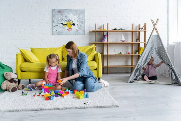 Kindergarten teacher playing building blocks with preschooler girl while kid with down syndrome sitting in tipi — Stock Photo