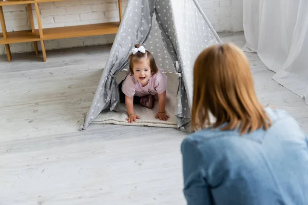 Blurred kindergarten teacher looking at happy kid with down syndrome sitting in tipi — Stock Photo