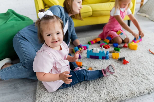 Niño pequeño con síndrome de Down mirando a la cámara mientras juega con la chica borrosa y el maestro de jardín de infantes en la alfombra - foto de stock