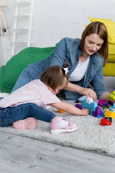 Toddler kid with down syndrome playing with smiling kindergarten teacher on carpet — Stock Photo